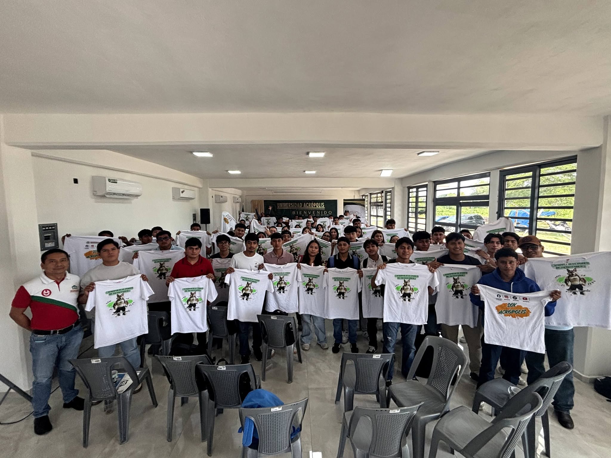 Estudiantes de nuevo ingreso de la modalidad no escolarizada sostienen playeras de la Universidad Acrópolis en un aula.