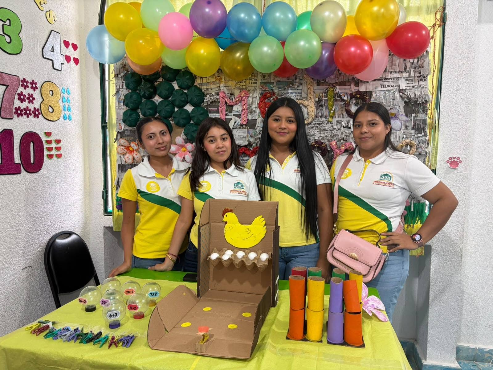 Cuatro estudiantes posan detrás de una mesa con materiales didácticos de cartón y un fondo decorado con globos.