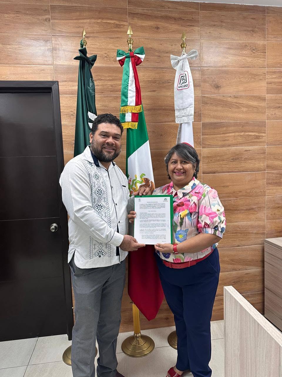 Un hombre y una mujer sostienen el convenio firmado frente a las banderas de México, el SPIUJAT y la Universidad Acrópolis.