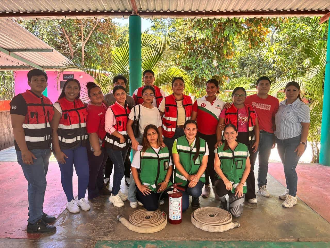 Participantes del taller posan con chalecos de seguridad detrás de un extintor y dos mangueras contra incendios enrolladas.